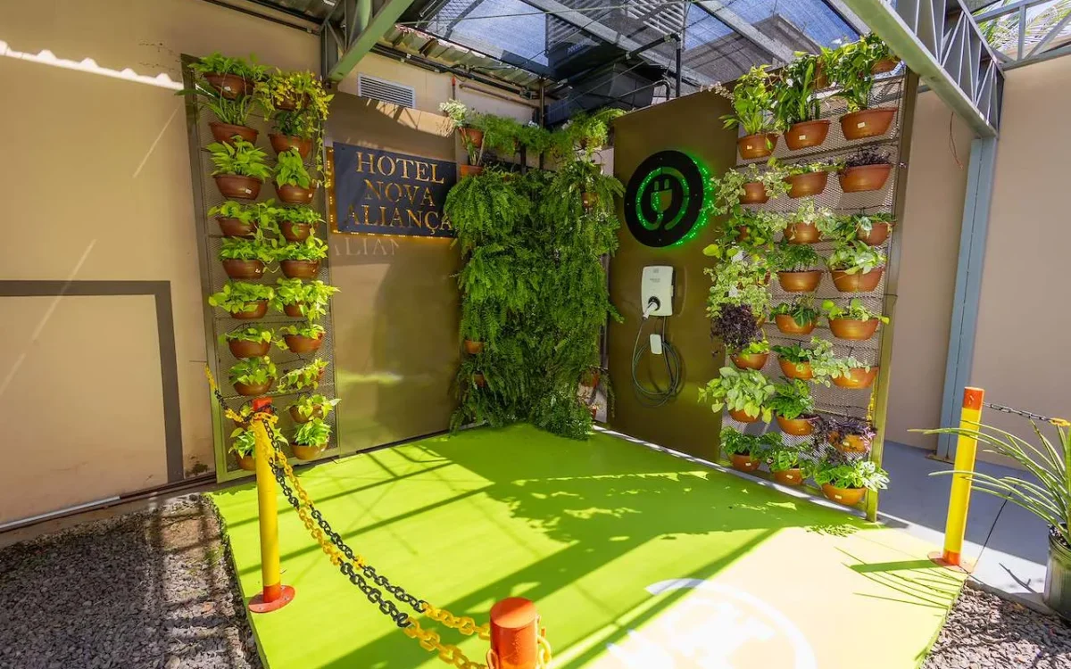 Espaço interior com paredes verdes, plantas em vasos de barro dispostas verticalmente, piso amarelo fluorescente, carregador de veículo elétrico na parede e pla