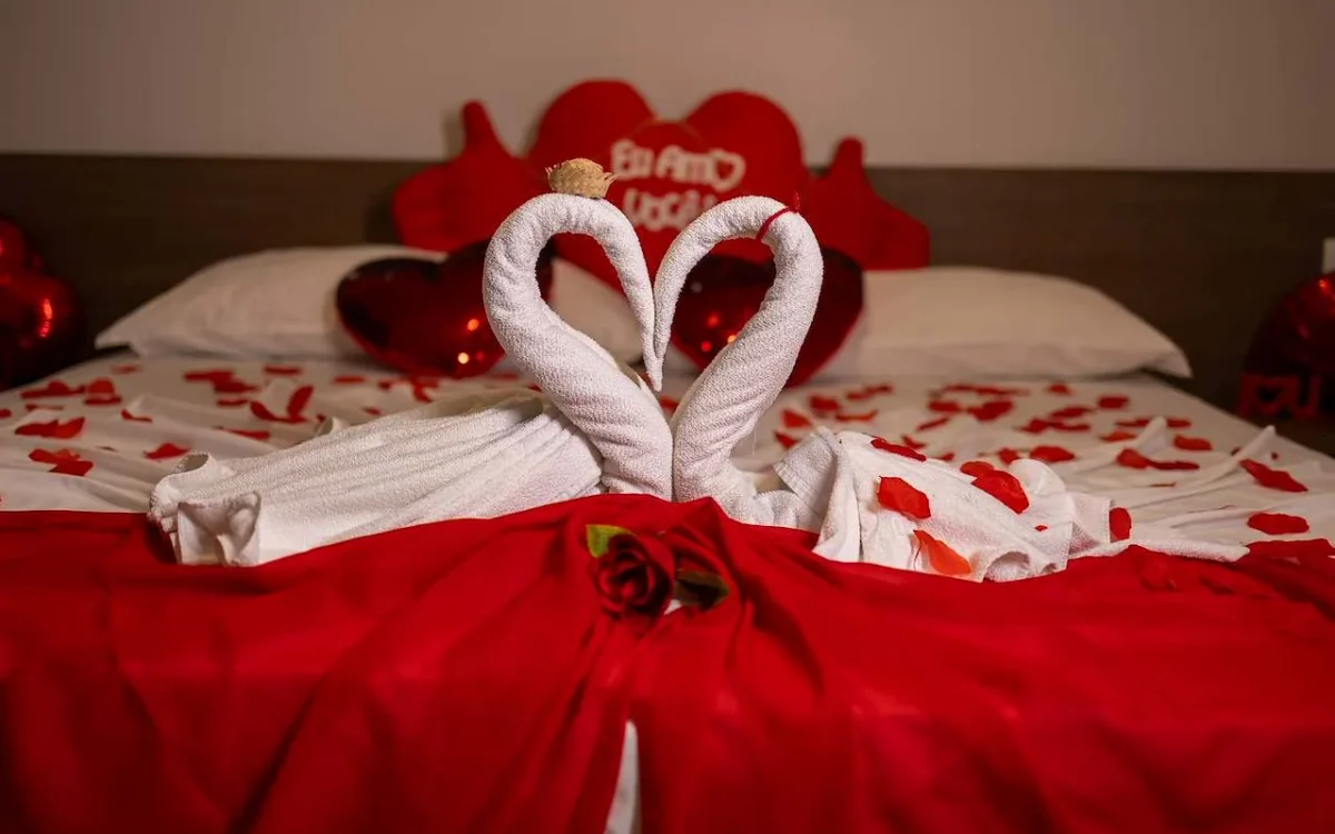 Duas toalhas brancas enroladas formando um coração sobre tecido vermelho com pétalas vermelhas e uma rosa