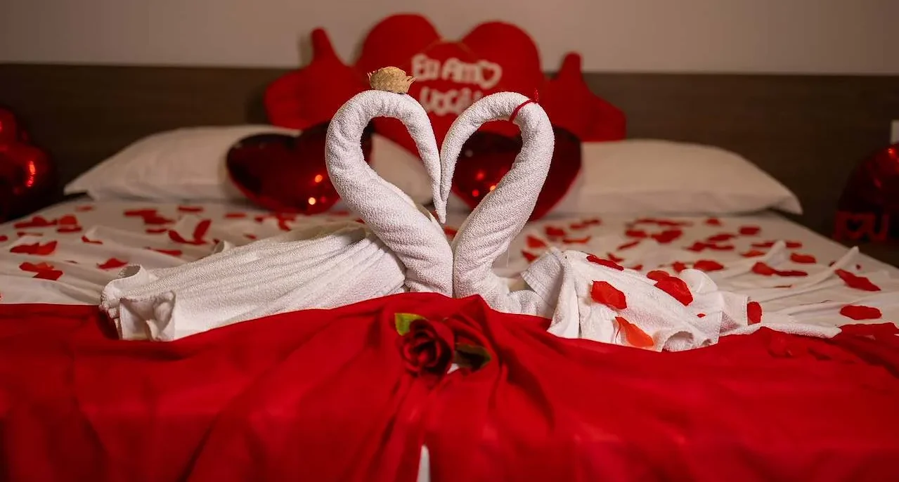 Duas toalhas brancas enroladas formando um coração sobre tecido vermelho com pétalas vermelhas e uma rosa