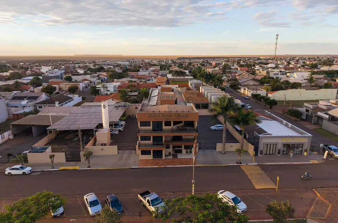 Vista aérea de uma cidade com hotel de três andares em primeiro plano, rua com carros estacionados, casas residenciais e céu parcialmente nublado ao fundo