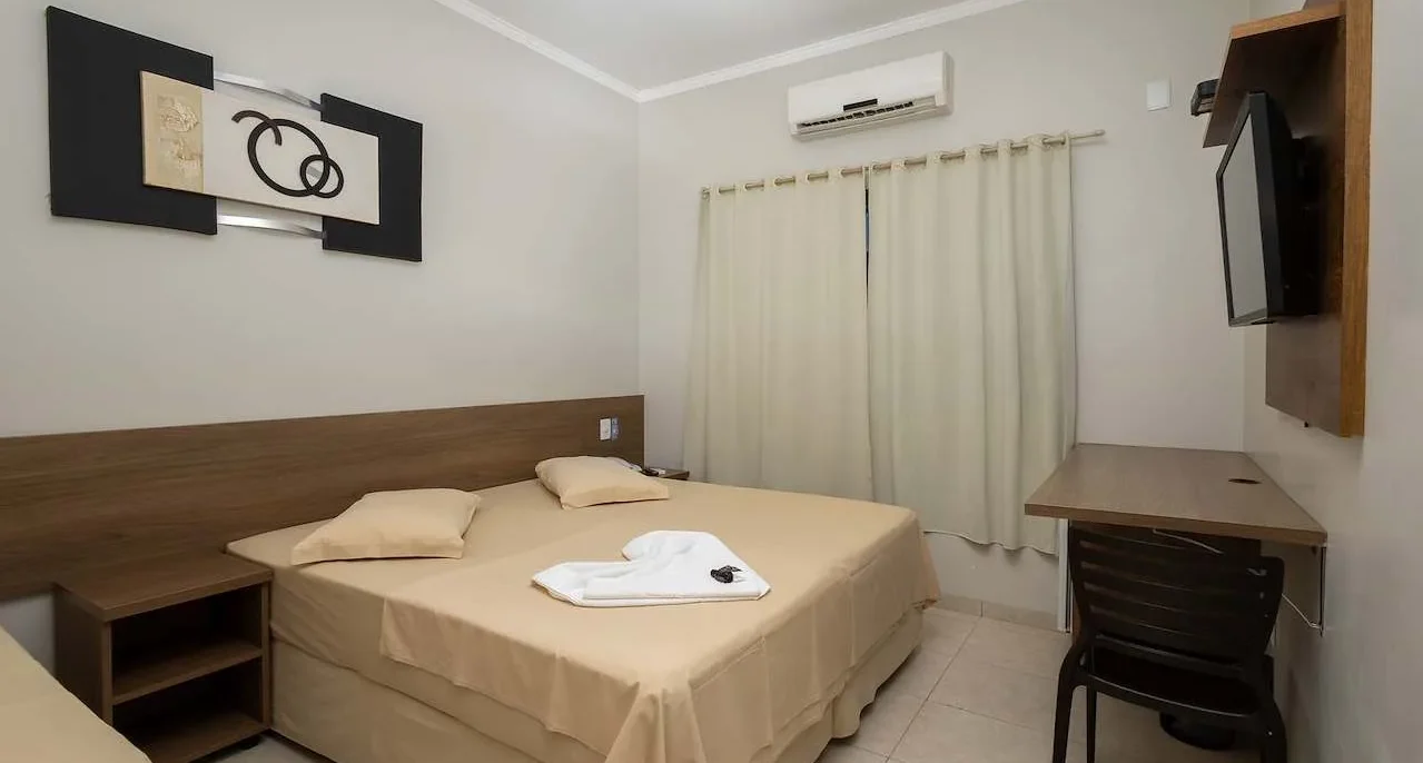 Quarto com cama de casal com roupa de cama bege, criado de madeira, cortinas claras, ar-condicionado, TV na parede e móvel de madeira