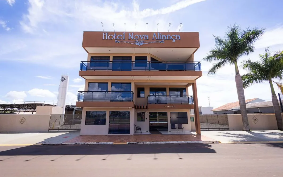 Fachada frontal de um edifício de quatro andares com nome Hotel Nova Aliança em letras azuis, palmeiras ao lado e céu azul ao fundo