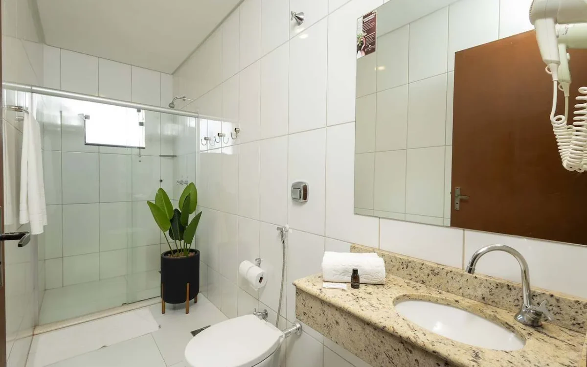 Banheiro moderno com azulejos brancos, espelho grande, pia de granito bege, vaso sanitário branco, chuveiro com box de vidro e planta decorativa preta