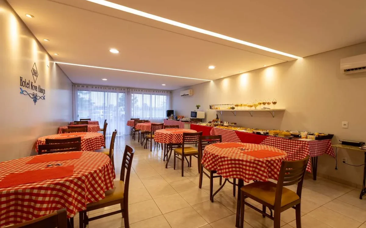 Sala de refeições com mesas redondas cobertas por toalhas vermelhas e brancas xadrez, cadeiras de madeira, luminária de teto moderna e balcão de serviço ao fund