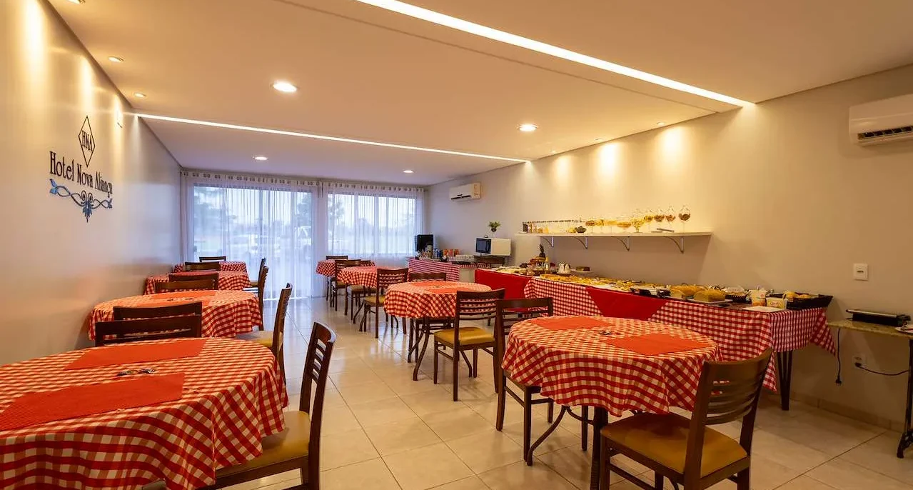 Sala de refeições com mesas redondas cobertas por toalhas vermelhas e brancas xadrez, cadeiras de madeira, luminária de teto moderna e balcão de serviço ao fund