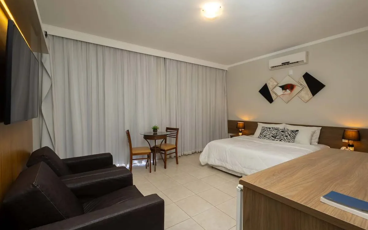 Quarto de hotel com cama de casal, sofá preto, mesa de trabalho, cortinas brancas e decoração moderna em tons neutros