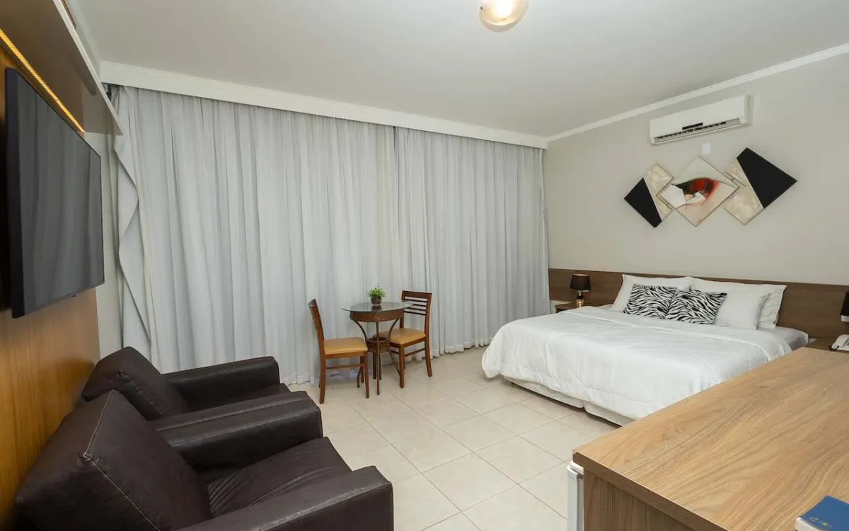 Quarto de hotel amplo com cama de casal, sofá preto, TV, cortinas brancas, mesa com cadeiras e ar-condicionado