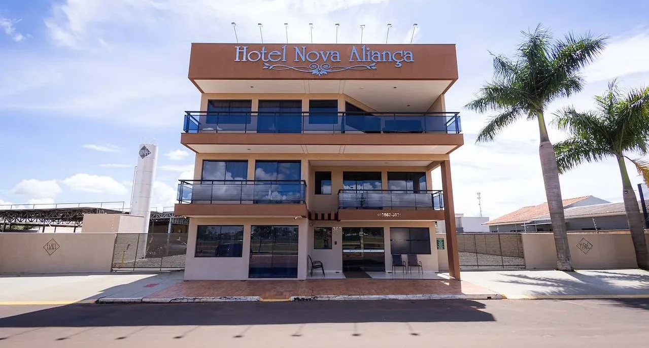 Fachada frontal de um edifício de quatro andares com nome Hotel Nova Aliança em letras azuis, palmeiras ao lado e céu azul ao fundo