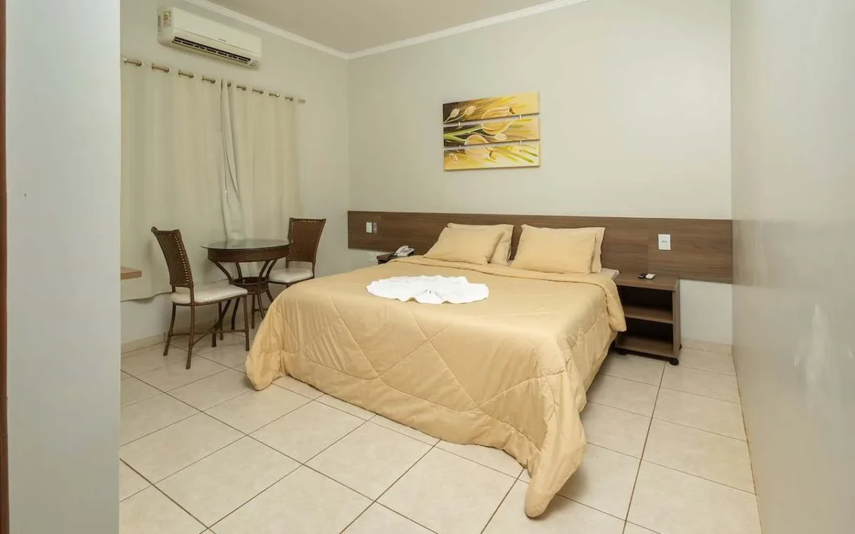 Quarto com cama de casal com roupa de cama bege, criado-mudo, mesa redonda e cadeiras, cortinas, ar-condicionado e quadro decorativo na parede