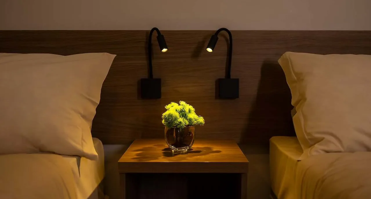 Dois sofás amarelos com almofadas, separados por mesa de madeira com vaso de flores amarelas, iluminados por abajures pretos na parede de madeira escura