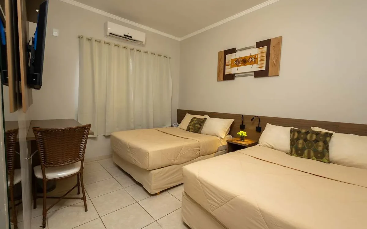 Quarto com dois leitos simples lado a lado, criado com abajur, quadro decorativo na parede, cortinas brancas, ar condicionado e cadeira de vime