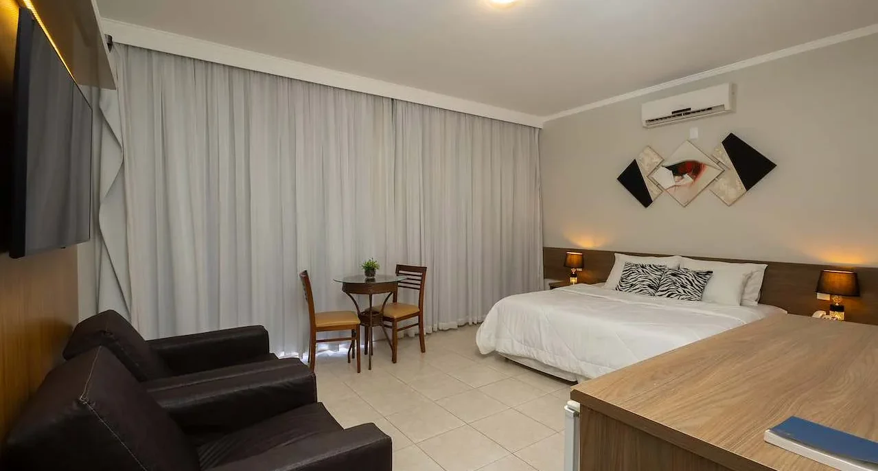 Quarto de hotel com cama de casal, sofá preto, mesa de trabalho, cortinas brancas e decoração moderna em tons neutros