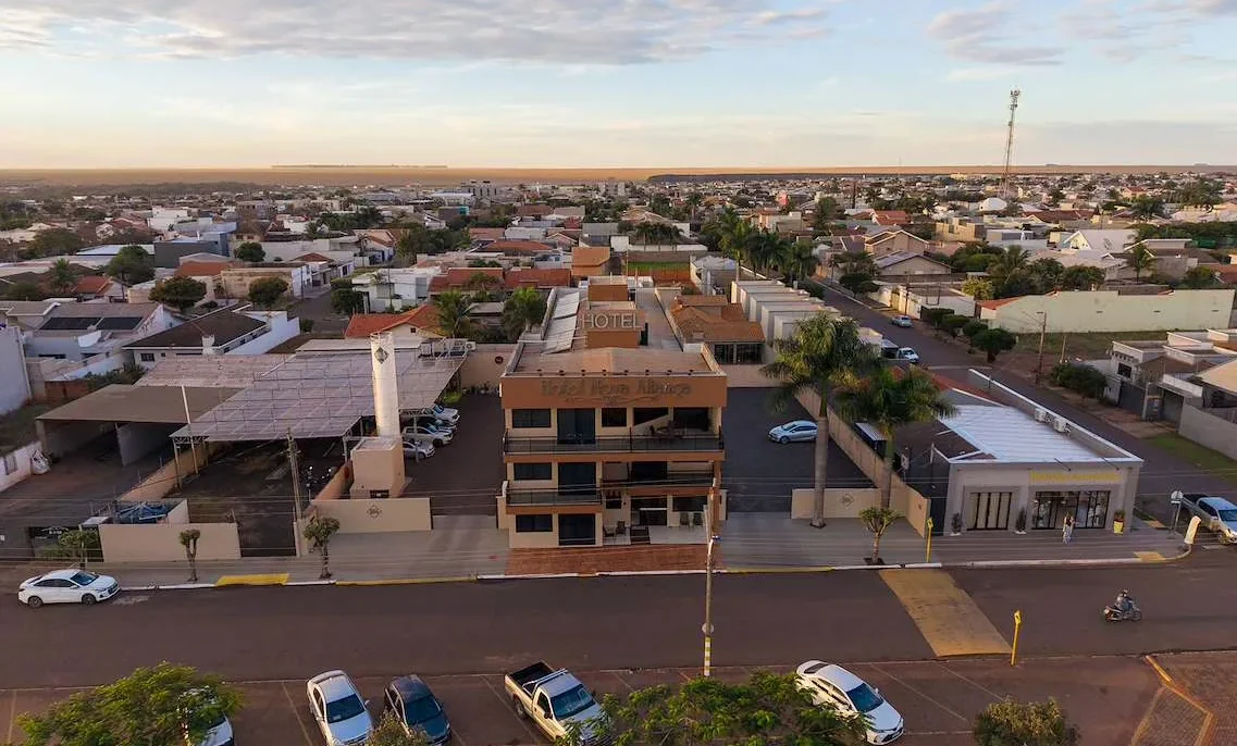 Vista aérea de uma cidade com hotel de três andares em primeiro plano, rua com carros estacionados, casas residenciais e céu parcialmente nublado ao fundo