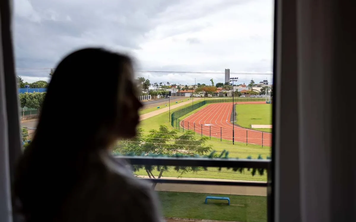 Pessoa com cabelo longo vista de costas olhando pela janela para um estádio de atletismo com pista vermelha e campos verdes