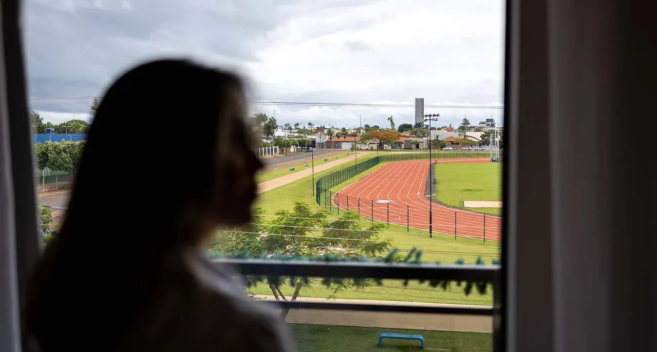 Pessoa com cabelo longo vista de costas olhando pela janela para um estádio de atletismo com pista vermelha e campos verdes