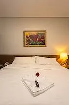 Cama com roupa de cama branca, duas almofadas, dois abajures acesos nas laterais, quadro na parede acima da cama e duas taças sobre a cama