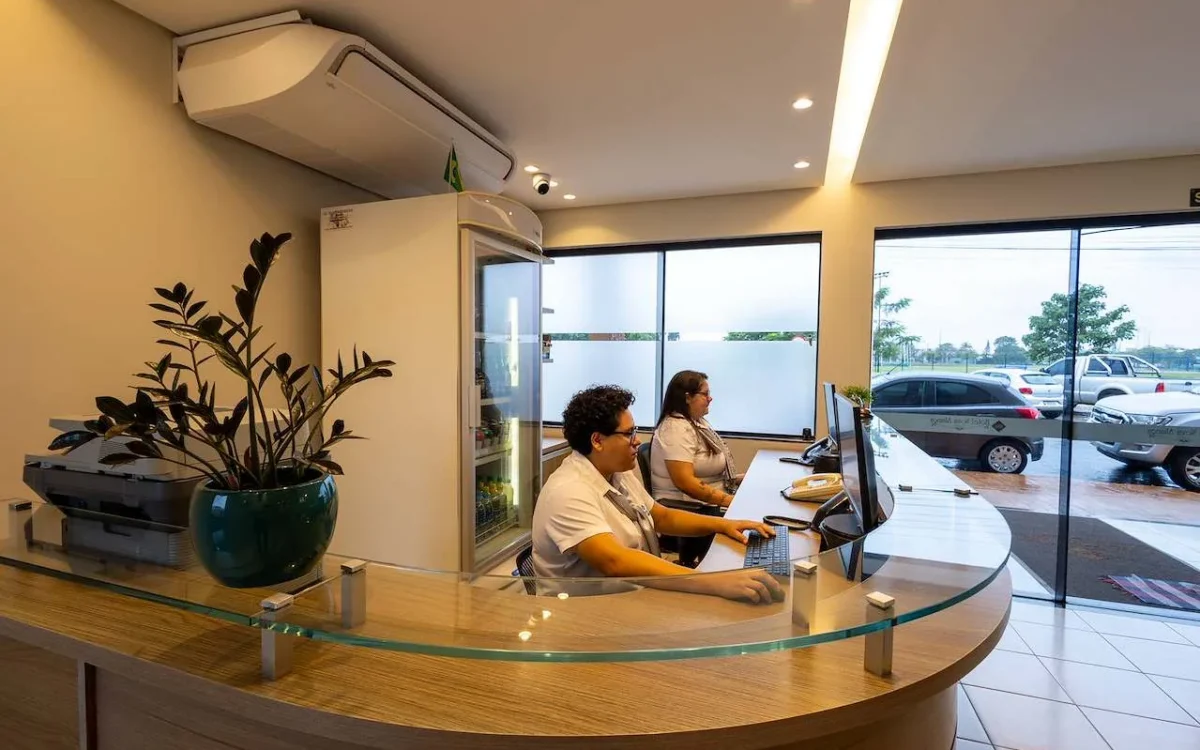 Dois funcionários trabalham em um balcão curvo com vidro em uma recepção moderna, com plantas, computadores e grandes janelas com vista para um estacionamento