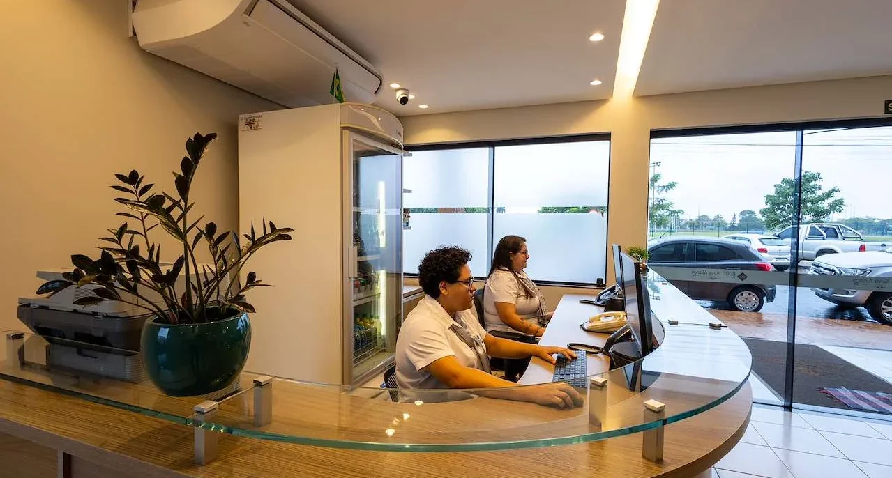 Dois funcionários trabalham em um balcão curvo com vidro em uma recepção moderna, com plantas, computadores e grandes janelas com vista para um estacionamento
