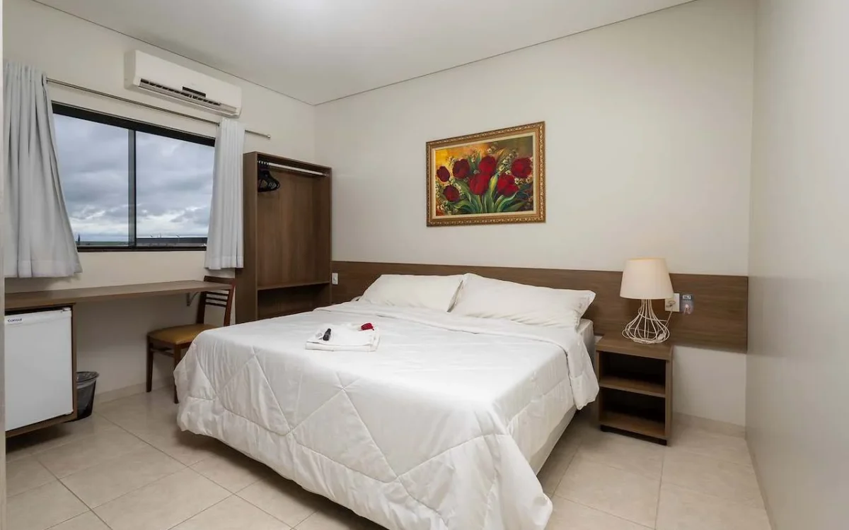 Quarto com cama de casal com roupa de cama branca, criado com luminária, janela com cortinas, ar-condicionado, quadro com flores vermelhas e móveis em tons de m
