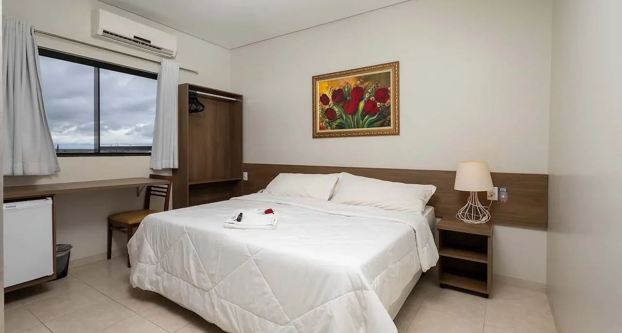 Quarto com cama de casal com roupa de cama branca, criado com luminária, janela com cortinas, ar-condicionado, quadro com flores vermelhas e móveis em tons de m