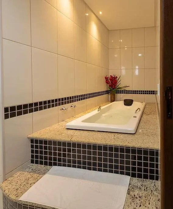 Banheiro moderno com banheira embutida em granito, azulejos brancos e pretos, espelho grande, iluminação em LED, flores vermelhas e toalha branca
