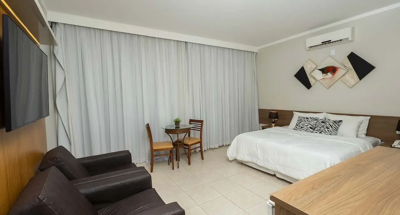 Quarto de hotel amplo com cama de casal, sofá preto, TV, cortinas brancas, mesa com cadeiras e ar-condicionado
