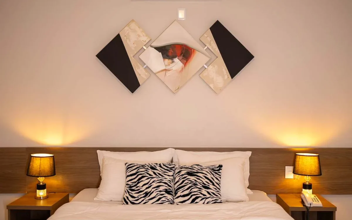 Quarto com cama de casal, almofadas estampadas em zebra, luminárias douradas nos criados, parede com obras de arte abstratas e iluminação ambient